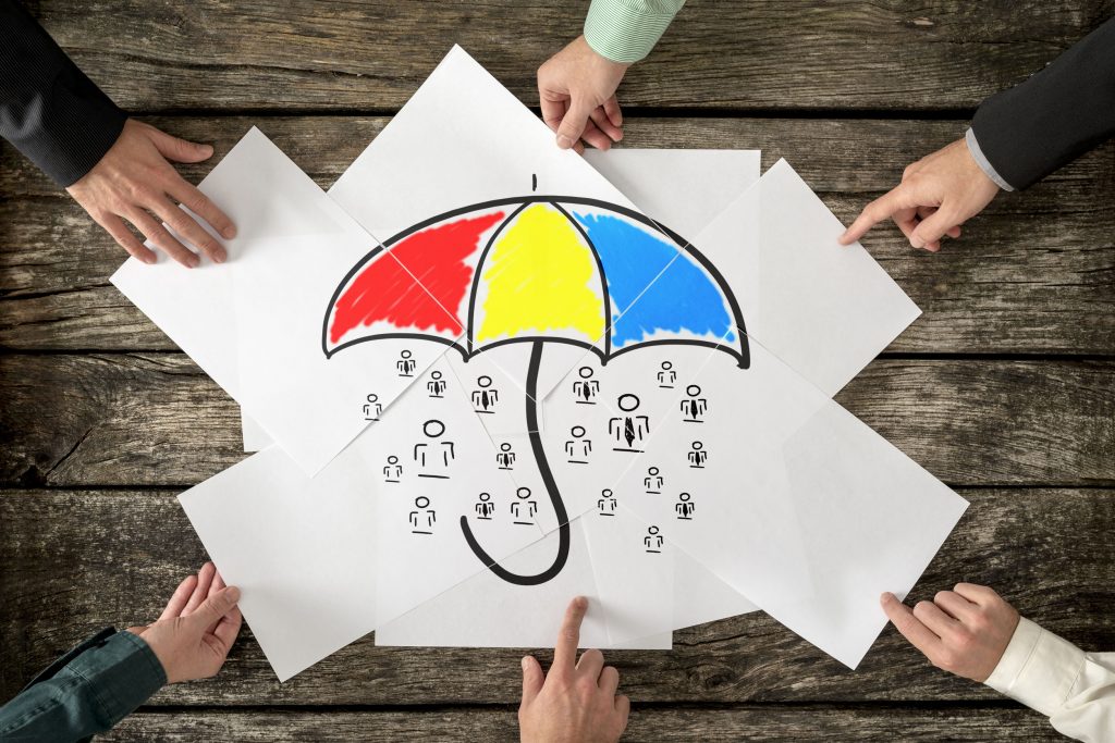 Safety and life insurance concept - six hands assembling a colourful umbrella sheltering many people icons drawn on white papers.