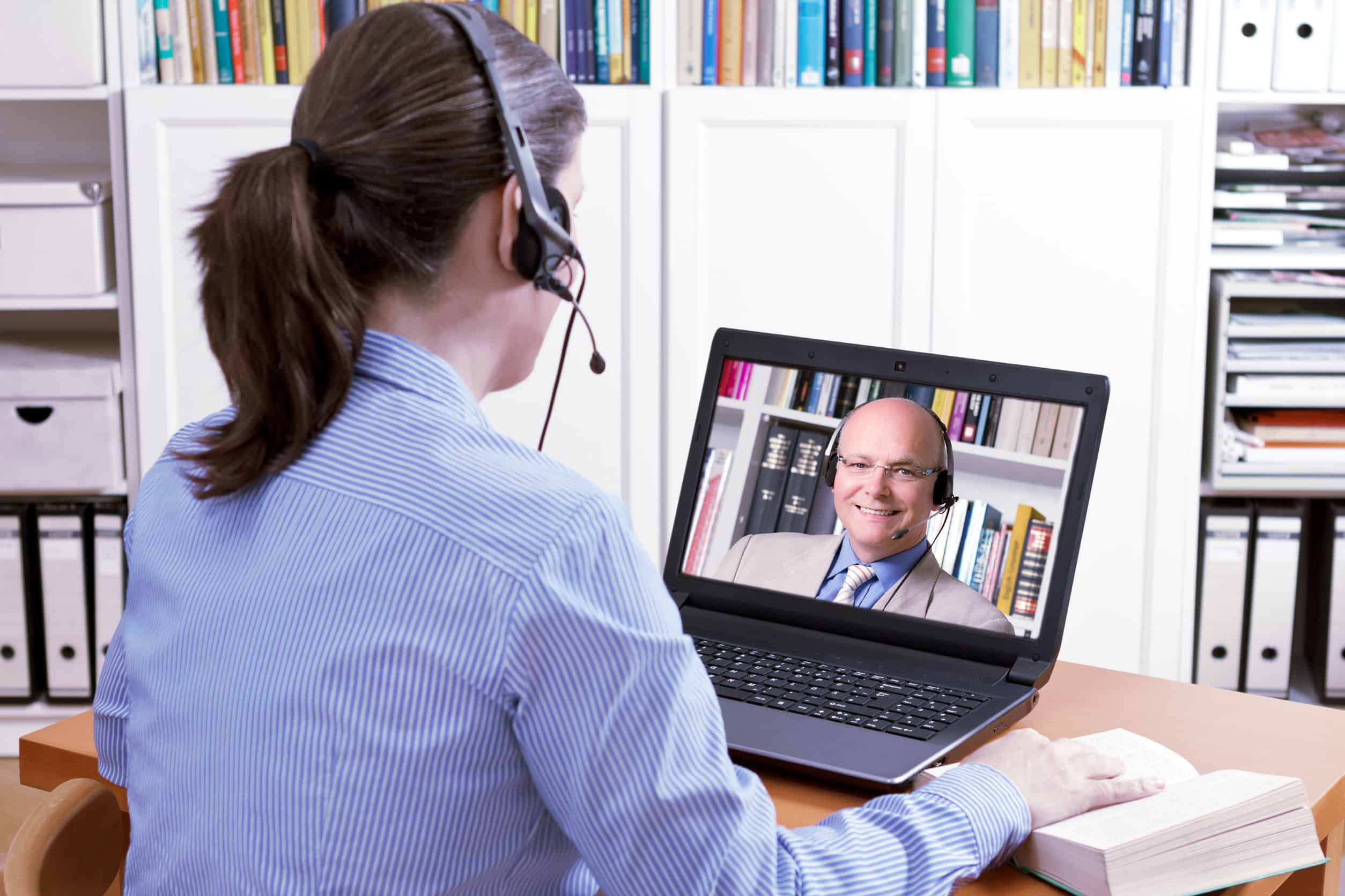 Woman with a headset in front of her laptop and a book making an online video call with her friendly teacher, text space, e-learning concept